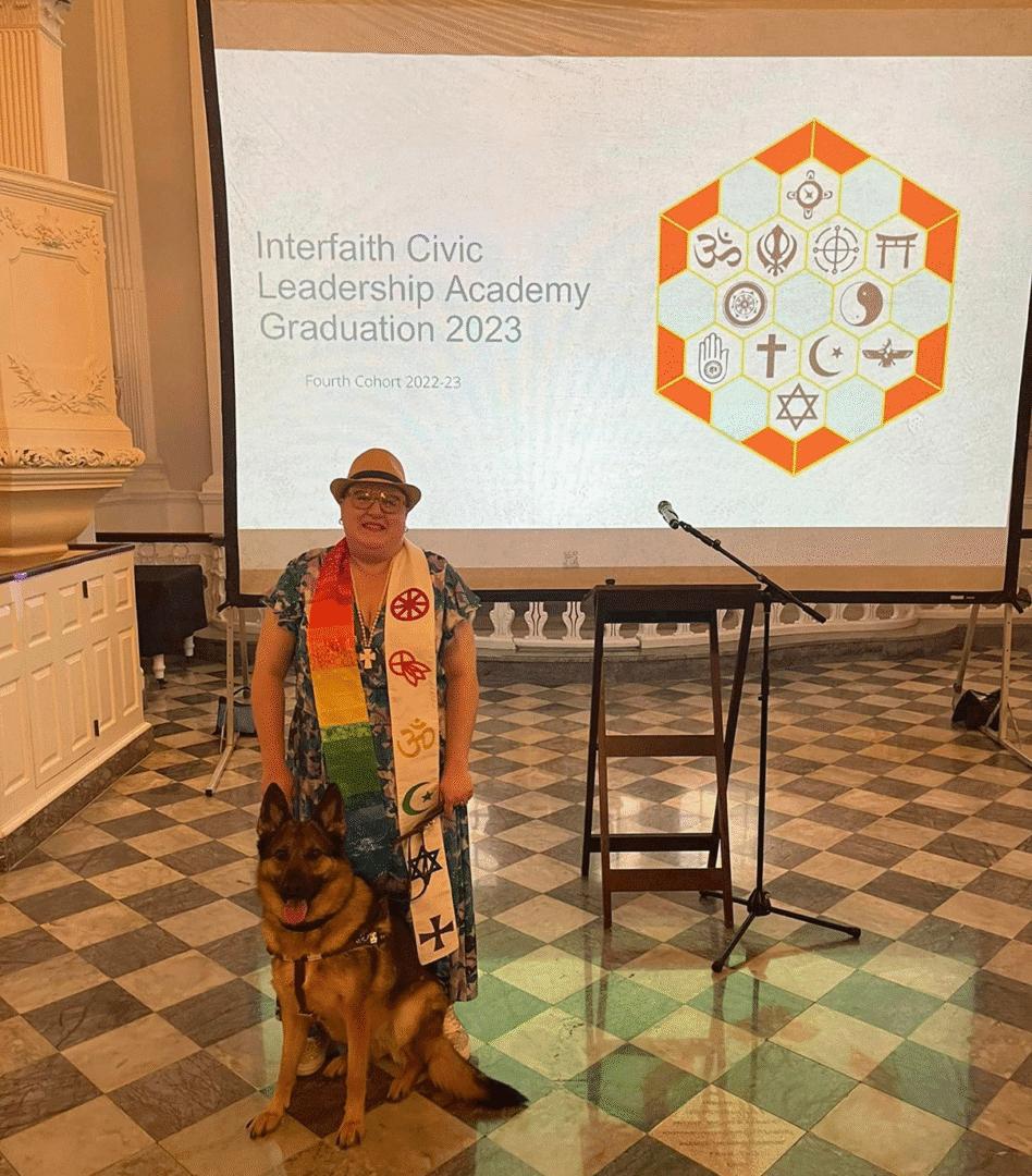 Dr. Bonner and her guide dog standing in front of a projector screen that reads "Interfaith Civic Leadership Academy Graduation 2023". The screen also has a graphic illustrating various faith group symbols.