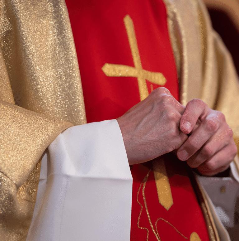 Closeup of a priest wearing a golden robe with a golden cross against red.