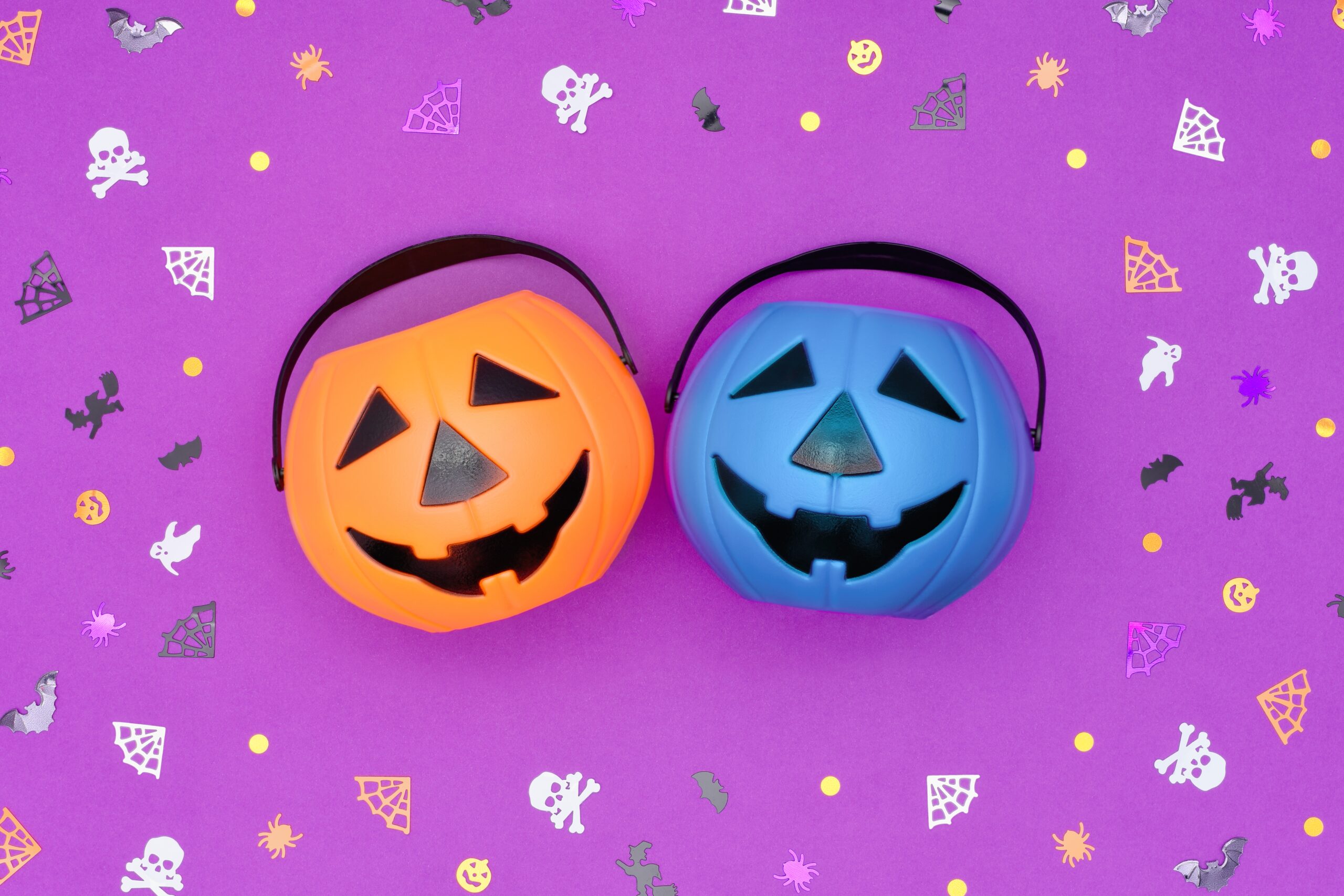 Two plastic pumpkin buckets—one orange and one teal blue—sit side by side on a purple background decorated with Halloween confetti, including bats, ghosts, spider webs, and skulls. The teal pumpkin represents allergy-friendly treats for trick-or-treaters.