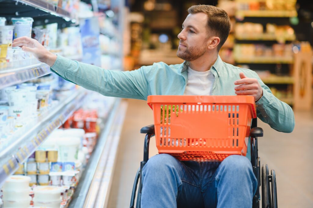 A man in a wheelchair shops in a grocery store, reaching for items on a refrigerated shelf while holding a shopping basket in his lap.