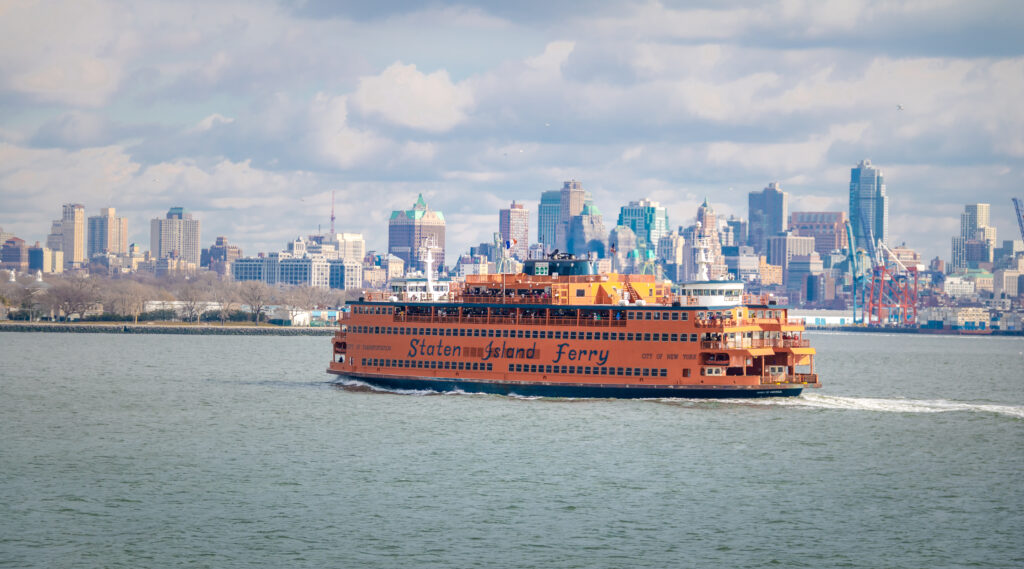 A bright orange Staten Island Ferry sails across New York Harbor with the Manhattan skyline in the background under a partly cloudy sky.