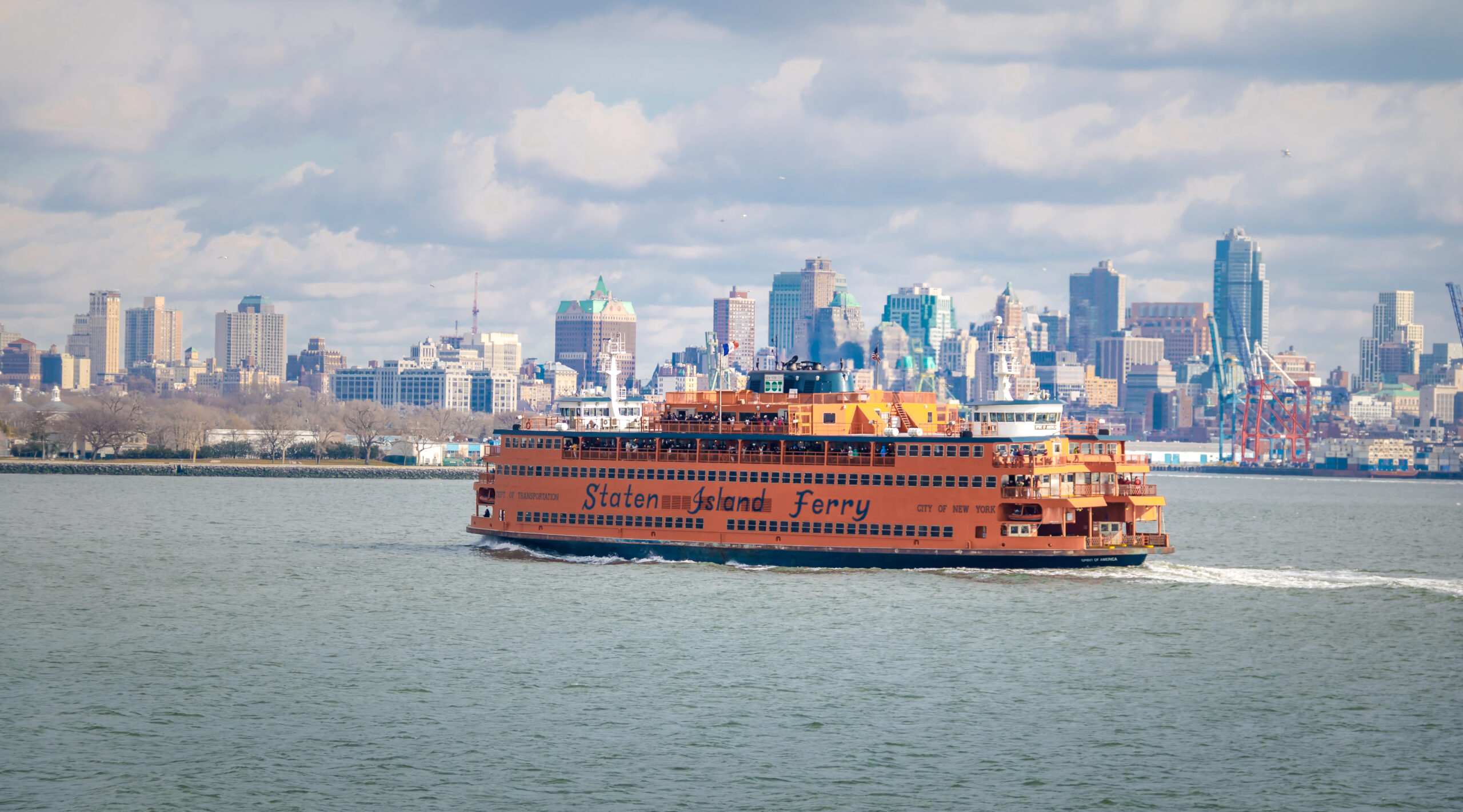 A bright orange Staten Island Ferry sails across New York Harbor with the Manhattan skyline in the background under a partly cloudy sky.