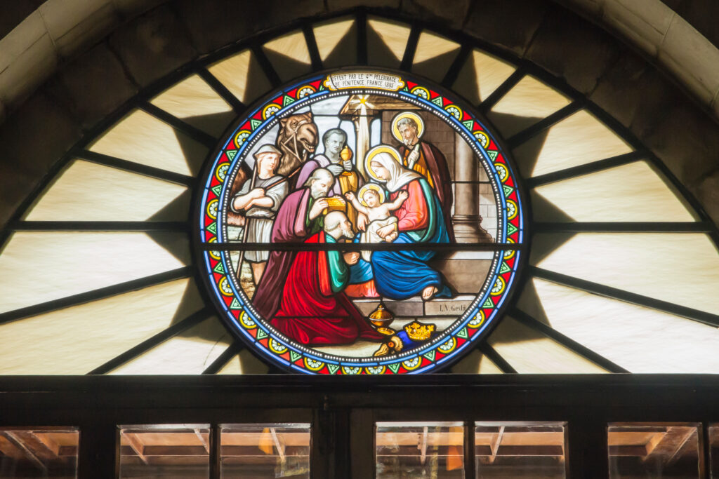 A circular stained glass window depicting the Nativity scene, showing the Virgin Mary holding baby Jesus as the three wise men present gifts, with Joseph and others looking on. The scene is framed by colorful geometric patterns, and a banner above reads “Offert par le Pèlerinage de Pénitence, France, 1885.”