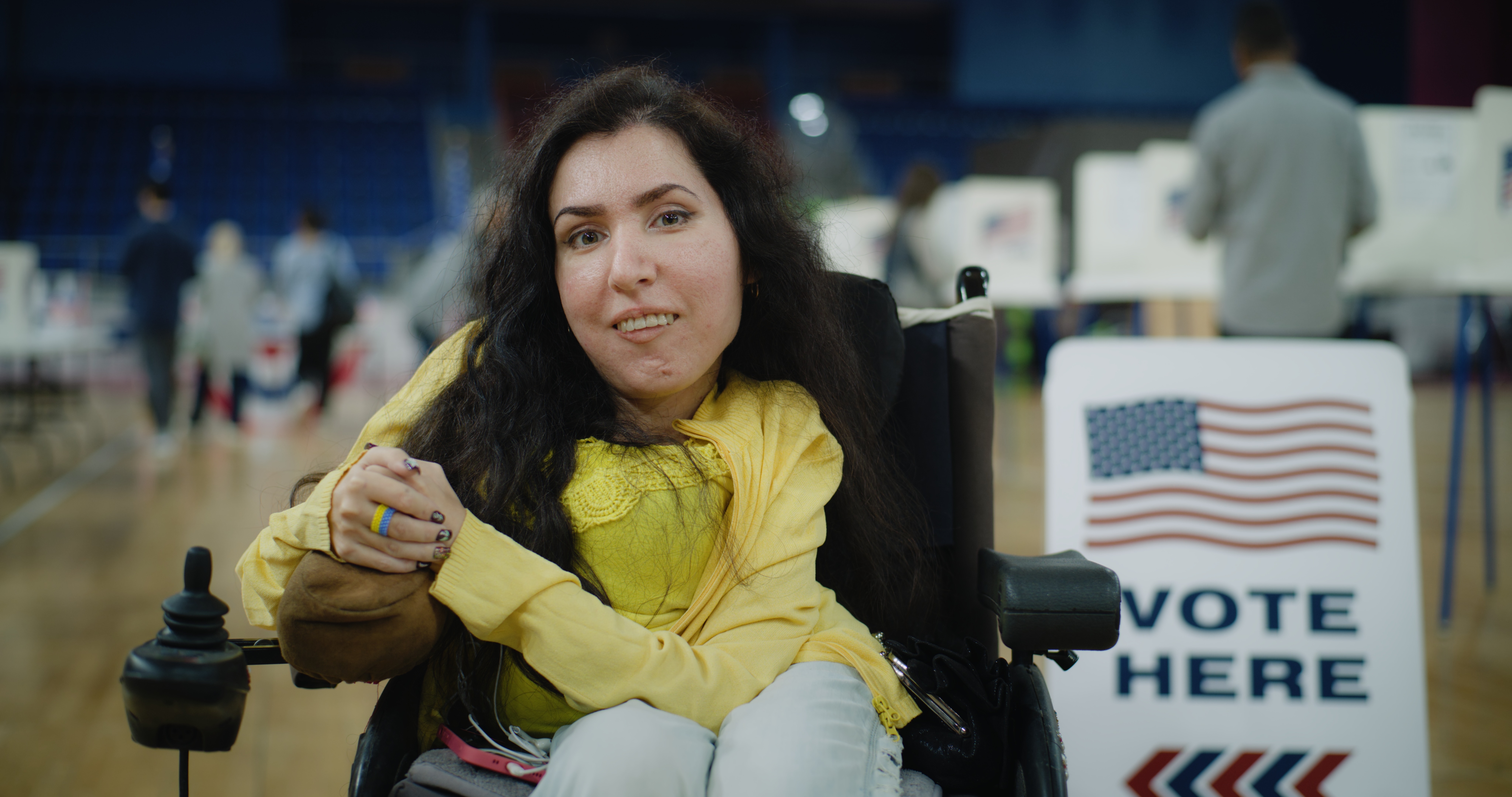 A woman in a wheelchair smiles at the camera inside a polling place. She is wearing a yellow top and sitting near a “Vote Here” sign decorated with an American flag. Other voters are blurred in the background as they cast their ballots.