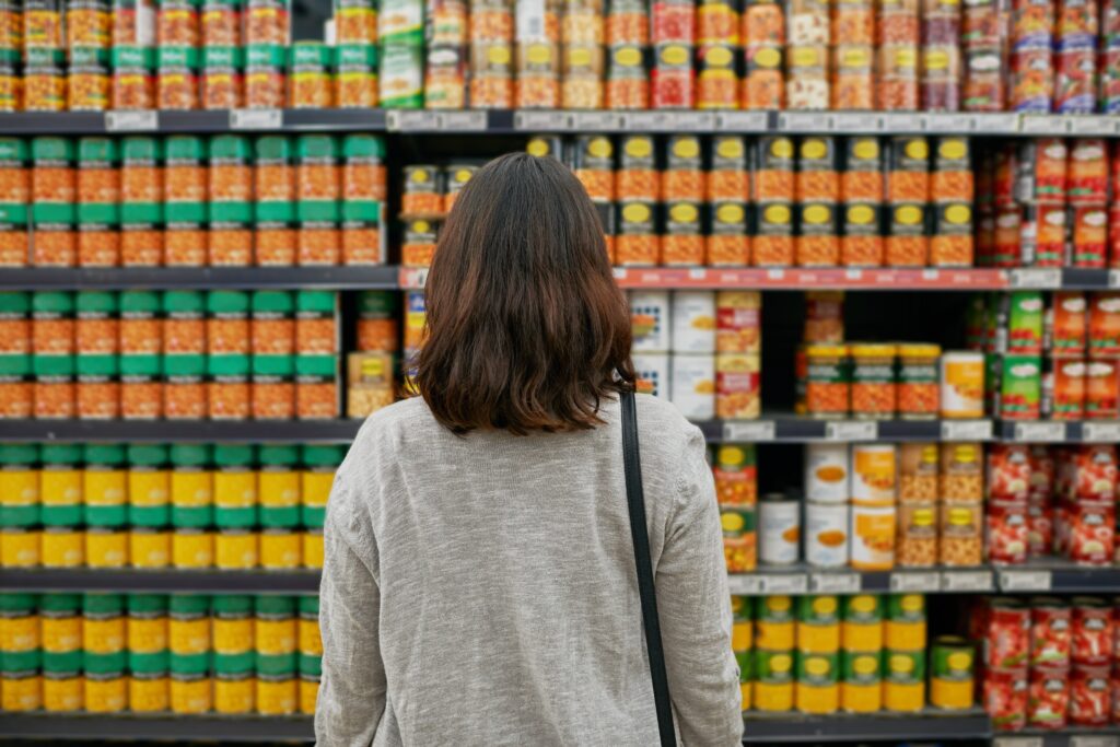 A woman stands in front of shelves of canned food, reflecting on the growing cost of groceries and the reality of hunger after SNAP benefits were cut.