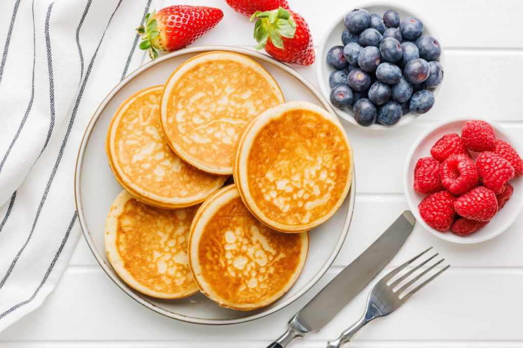 Overhead view of a plate stacked with golden-brown pancakes on a white table, surrounded by bowls of fresh blueberries and raspberries, whole strawberries, a fork and knife, and a white striped napkin.
