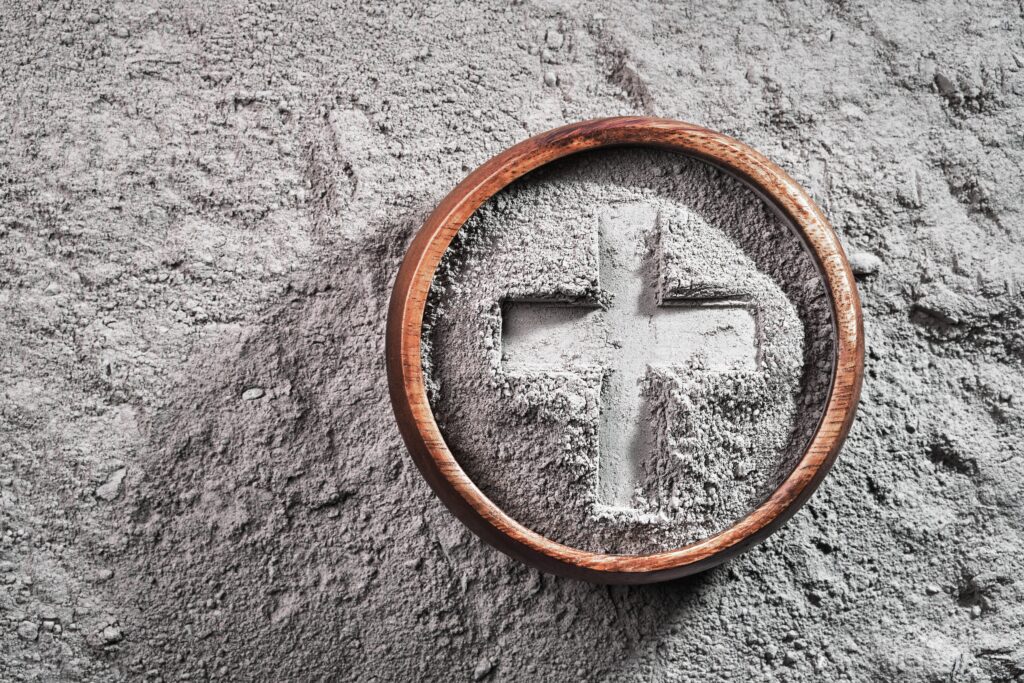 Close-up of gray ash arranged in the shape of a cross inside a round wooden bowl, resting on a textured gray surface.