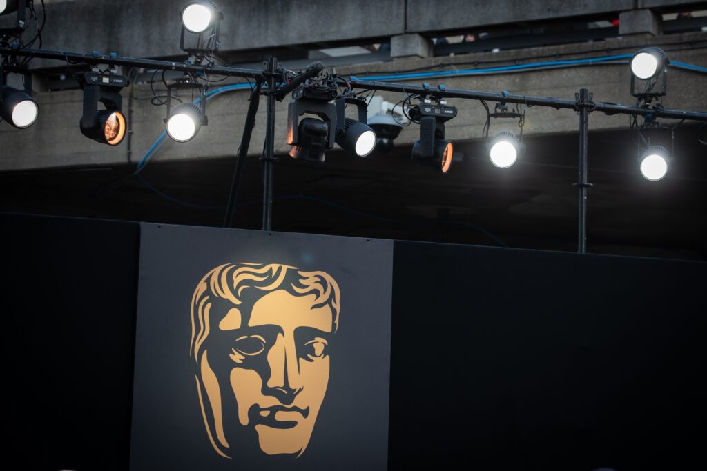 Stage lighting rig suspended above a black backdrop featuring the large gold BAFTA mask logo, illuminated by bright white spotlights during an awards ceremony.
