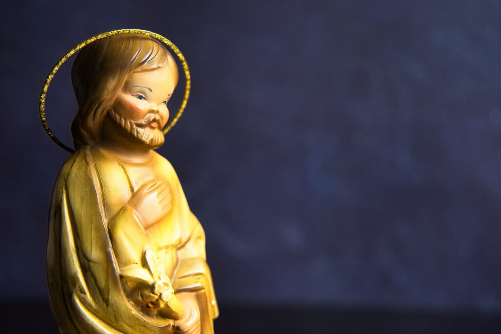 A small golden statue of Saint Joseph with a halo, shown in profile against a dark blue background, holding a lily and looking downward with a gentle expression.