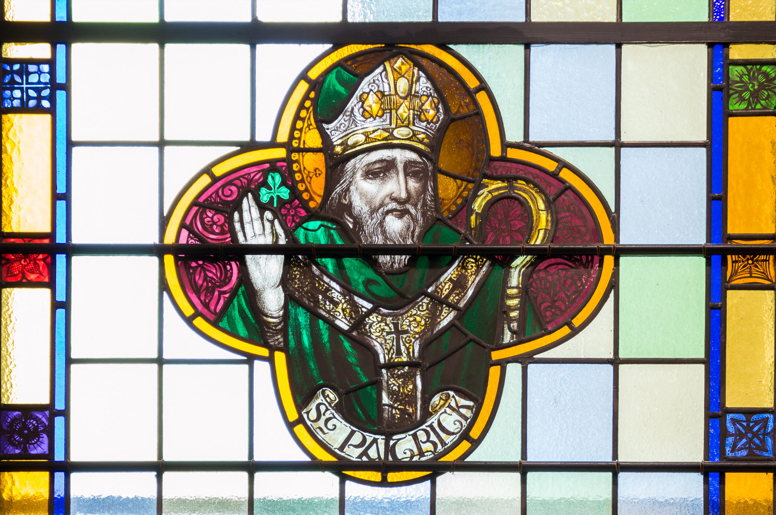 Stained glass window depicting Saint Patrick, shown wearing a bishop’s mitre and green robes, raising one hand in blessing while holding a crozier, framed by a clover-shaped design and colorful geometric panes in the background.