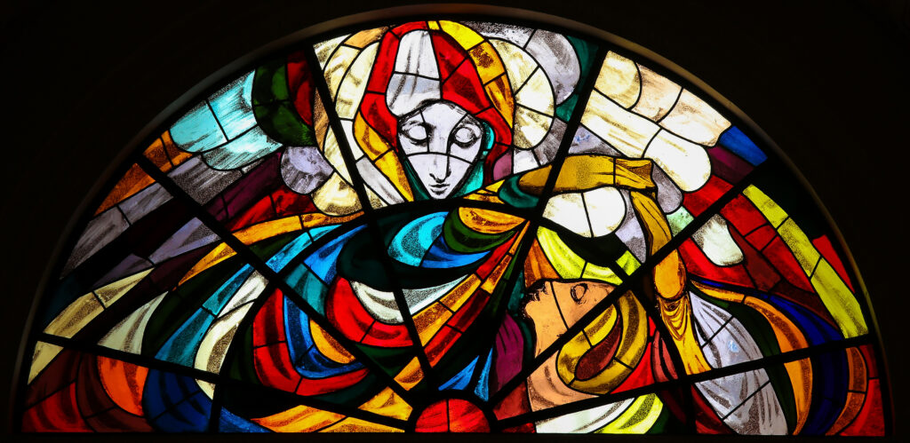 A colorful stained-glass window depicting a serene, Blessed Mother in flowing robes leaning toward another person who gazes upward, bathed in radiant light, with vivid reds, blues, yellows, and greens forming an abstract, sacred scene suggestive of a biblical encounter.