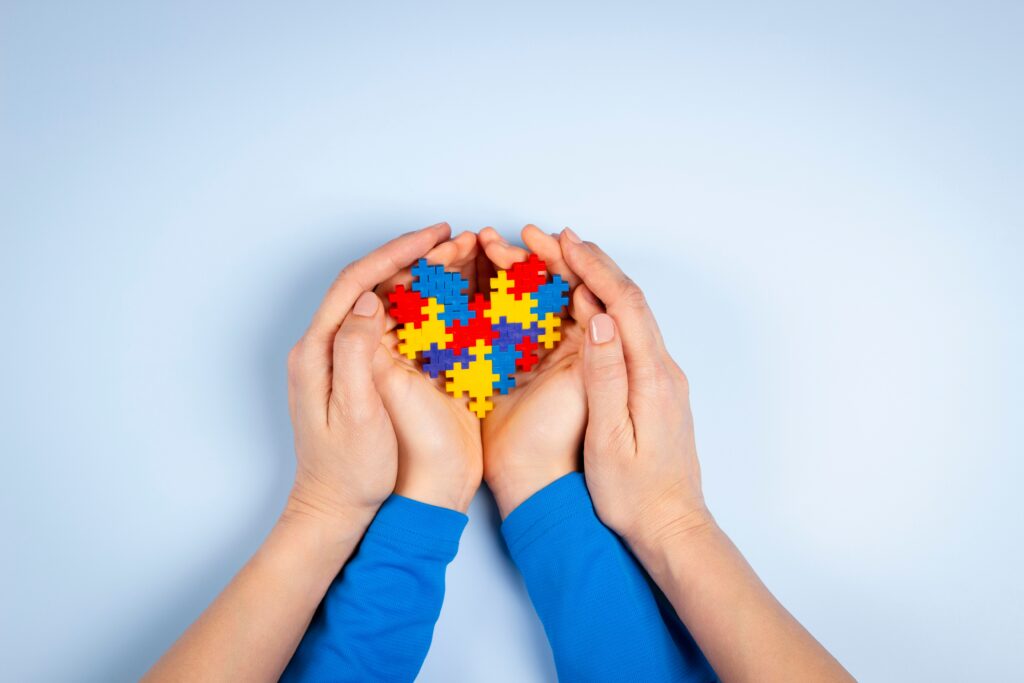 Two adult hands gently cup a child’s hands holding a heart made of colorful puzzle pieces, symbolizing love, care, and support for autism and neurodiversity.