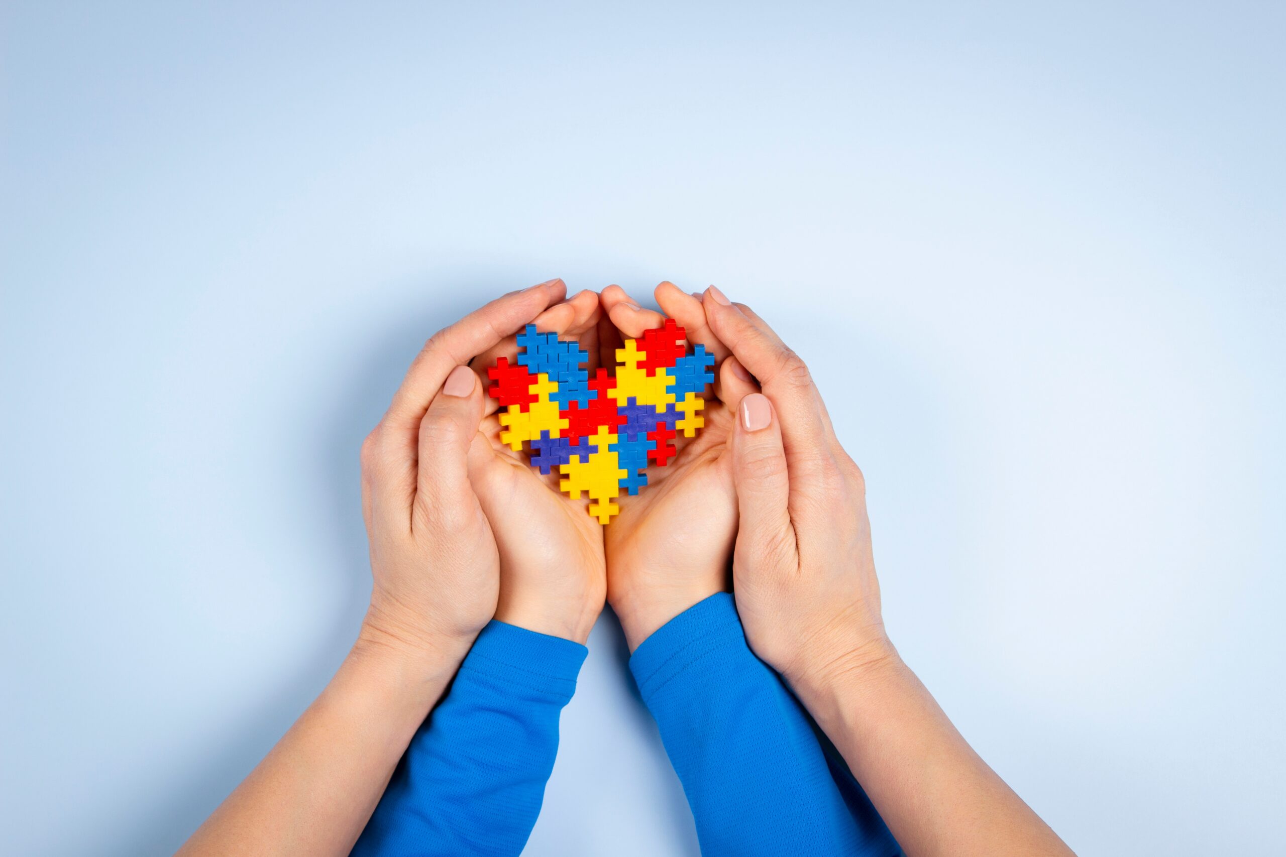 Two adult hands gently cup a child’s hands holding a heart made of colorful puzzle pieces, symbolizing love, care, and support for autism and neurodiversity.