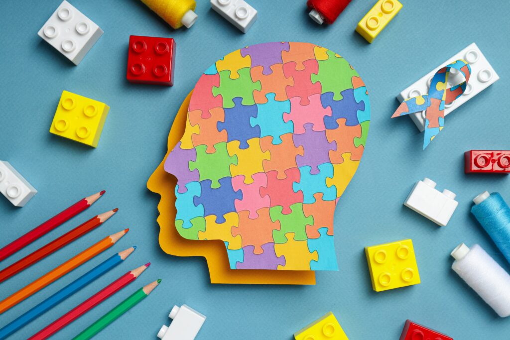 A colorful profile of a human head made of puzzle pieces sits on a blue background, surrounded by crayons, building blocks, and thread, symbolizing autism, creativity, and different ways of thinking.