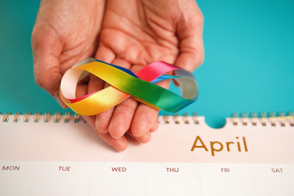 A hand holds a rainbow infinity ribbon above a calendar page labeled April, representing Autism Awareness Month and the beauty of neurodiversity.