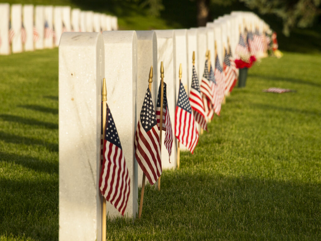 A peaceful Memorial Day cemetery scene stretches into the distance, with rows of white marble military headstones standing in neat formation across a green lawn. Small American flags are placed beside many of the graves, gently catching the light of the sun. The background is softly blurred, emphasizing the solemn beauty and quiet honor of the memorial setting. The image conveys remembrance, sacrifice, patriotism, and respect for fallen service members.