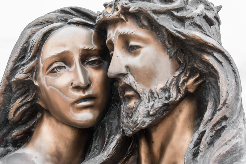 Close-up bronze sculpture of Jesus and Mary with their faces pressed closely together. Mary looks outward with a sorrowful expression, while Jesus, wearing a crown of thorns, tilts his head toward her. The sculpture has smooth polished highlights and textured details in the hair and beard against a light background.