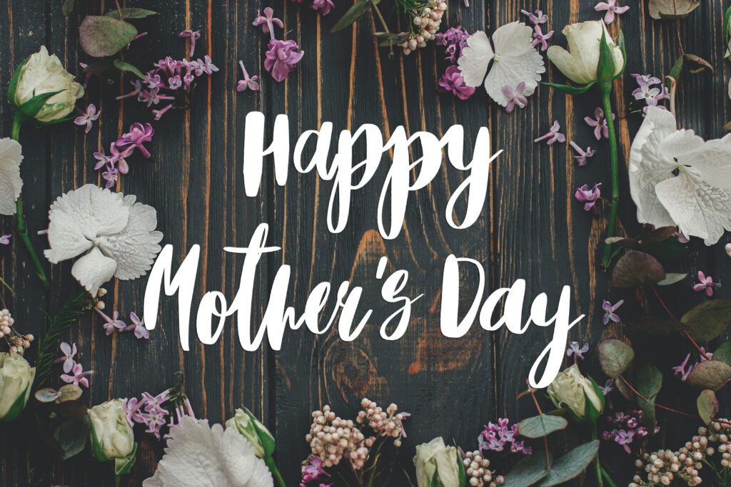 Decorative Mother’s Day graphic featuring white script text reading ‘Happy Mother’s Day’ centered on a dark rustic wooden background, surrounded by white flowers, purple lilacs, and green leaves.