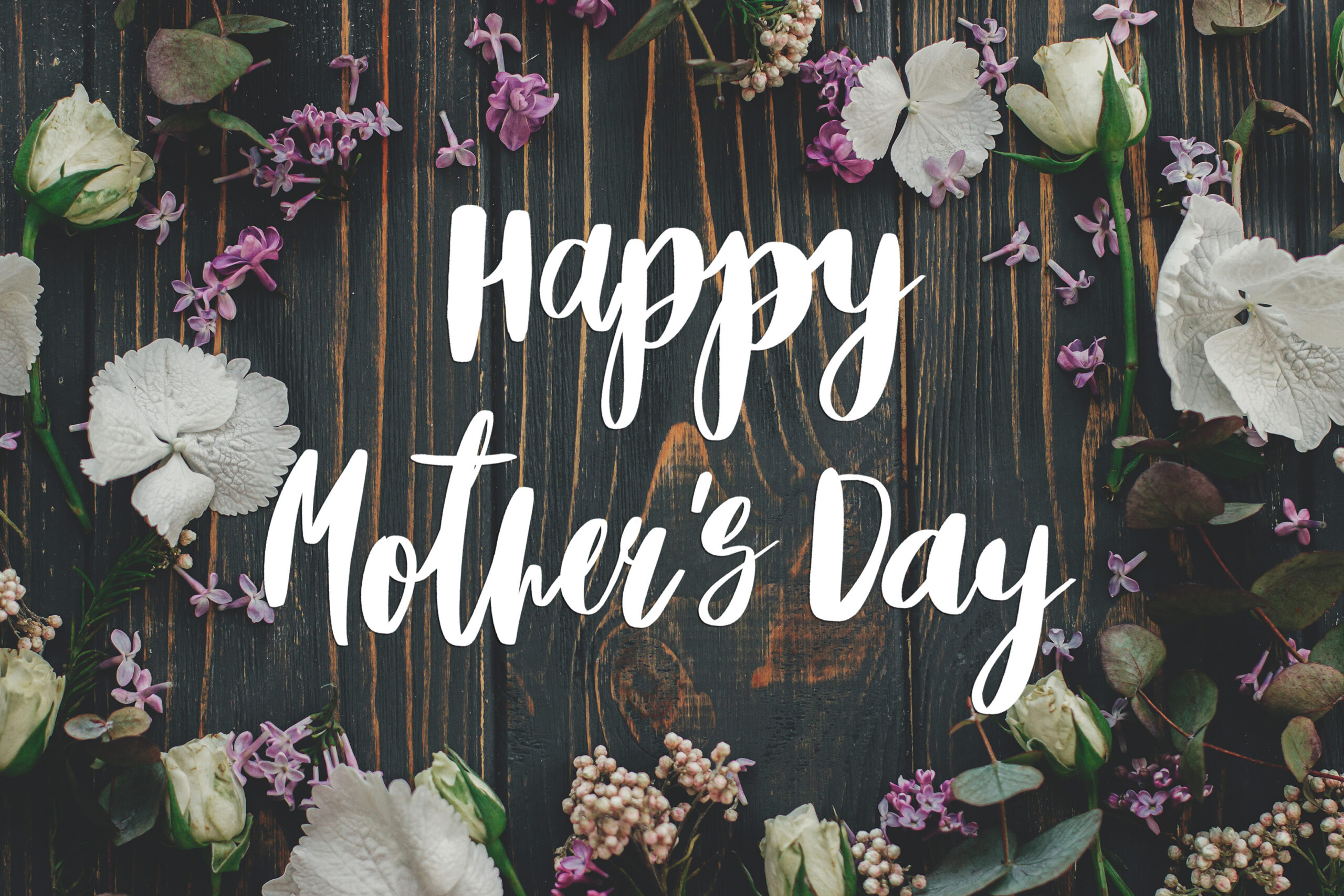 Decorative Mother’s Day graphic featuring white script text reading ‘Happy Mother’s Day’ centered on a dark rustic wooden background, surrounded by white flowers, purple lilacs, and green leaves.