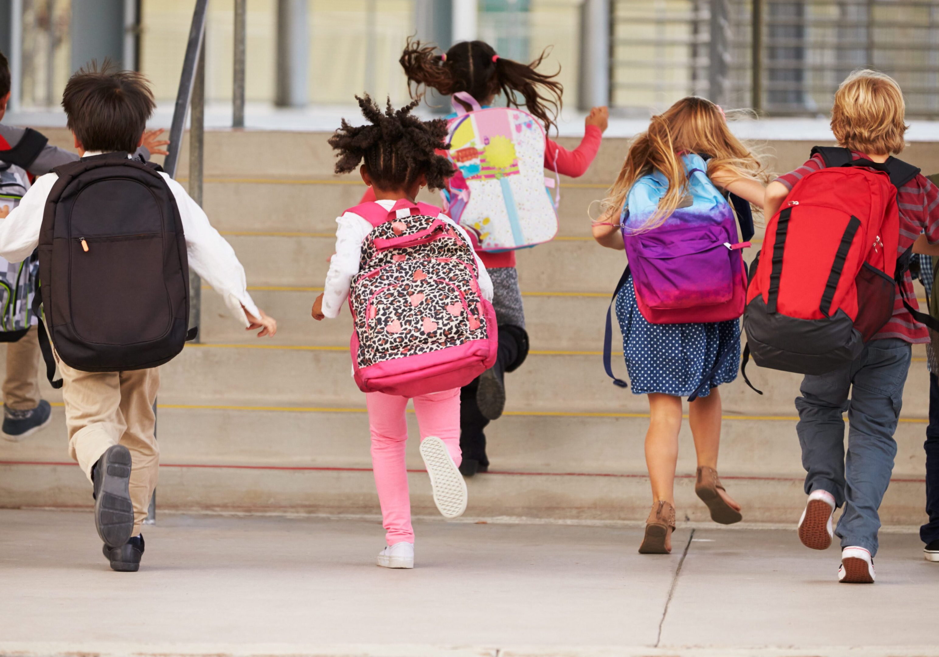 Group of children wearing backpacks, running up school steps together.