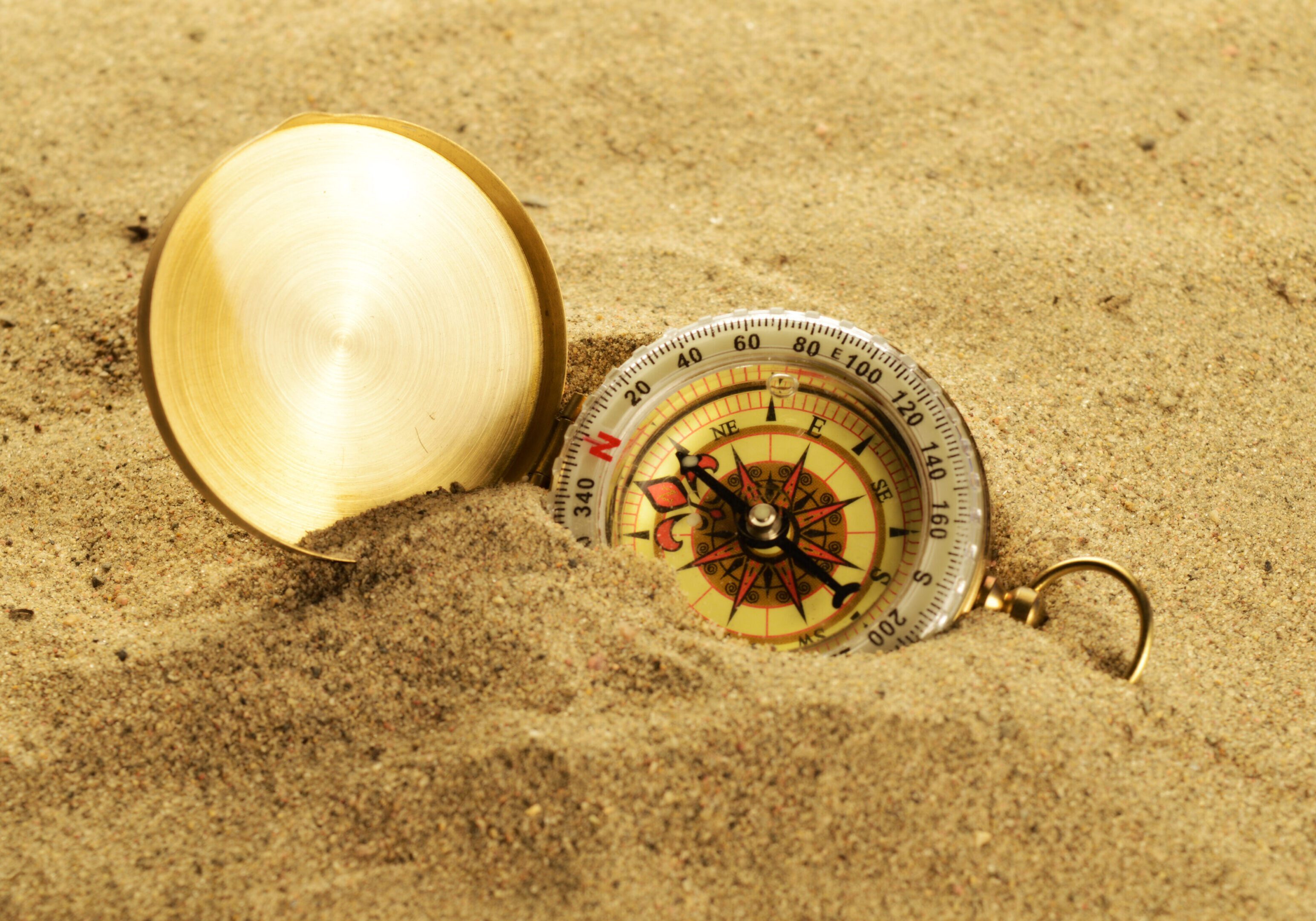 Compass half-buried in sand with its golden lid open beside it.