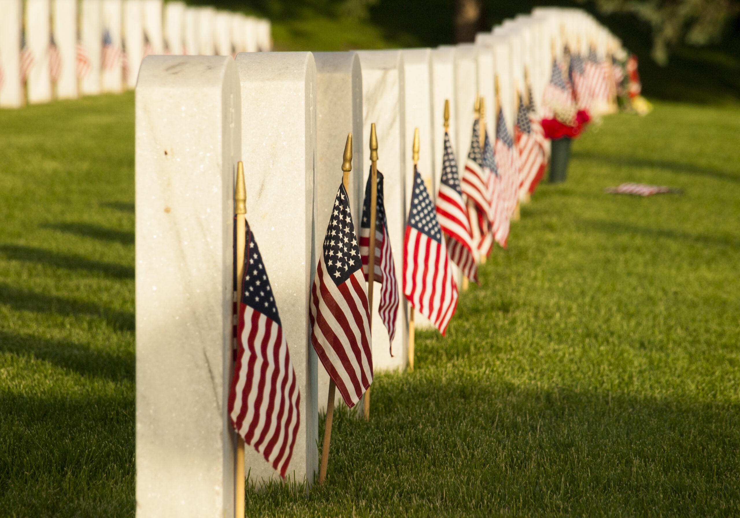 A peaceful Memorial Day cemetery scene stretches into the distance, with rows of white marble military headstones standing in neat formation across a green lawn. Small American flags are placed beside many of the graves, gently catching the light of the sun. The background is softly blurred, emphasizing the solemn beauty and quiet honor of the memorial setting. The image conveys remembrance, sacrifice, patriotism, and respect for fallen service members.