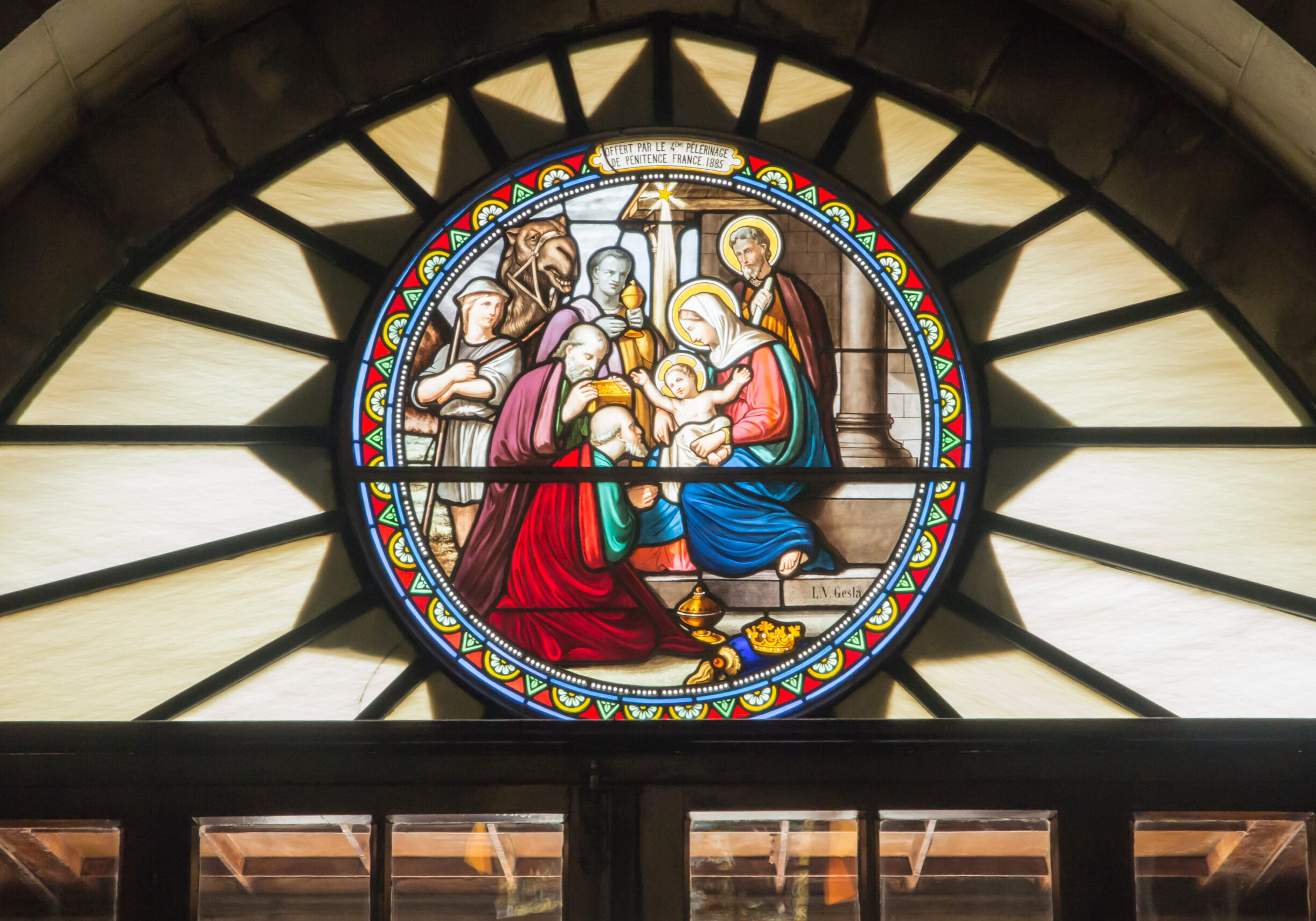 A circular stained glass window depicting the Nativity scene, showing the Virgin Mary holding baby Jesus as the three wise men present gifts, with Joseph and others looking on. The scene is framed by colorful geometric patterns, and a banner above reads “Offert par le Pèlerinage de Pénitence, France, 1885.”