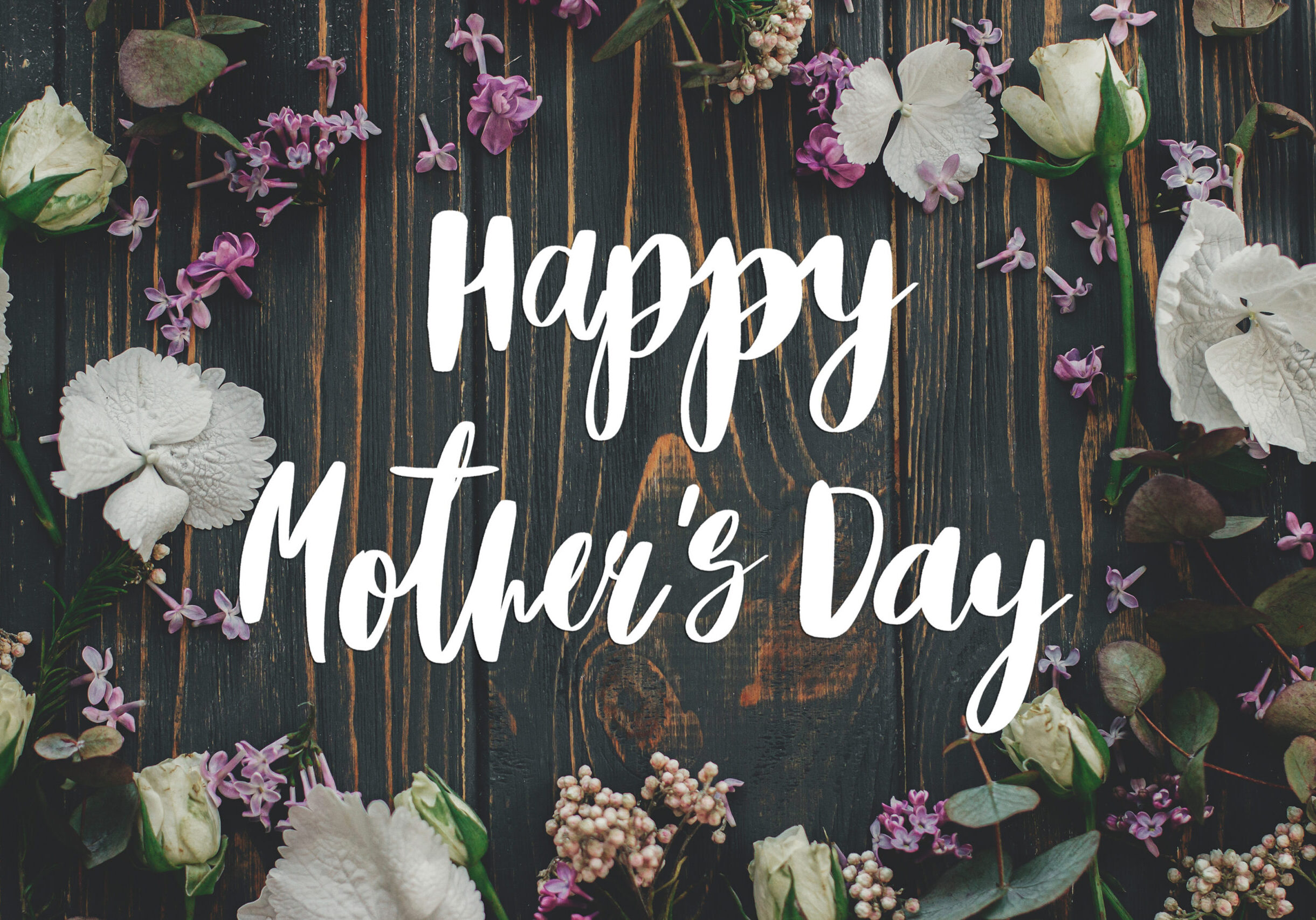 Decorative Mother’s Day graphic featuring white script text reading ‘Happy Mother’s Day’ centered on a dark rustic wooden background, surrounded by white flowers, purple lilacs, and green leaves.