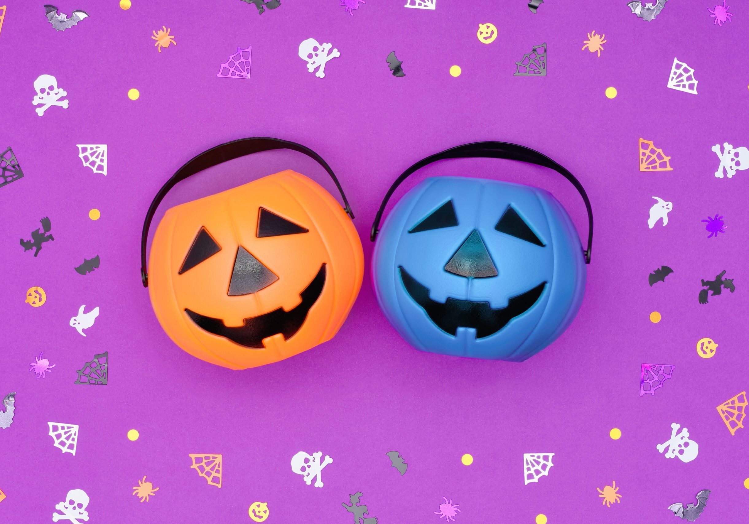 Two plastic pumpkin buckets—one orange and one teal blue—sit side by side on a purple background decorated with Halloween confetti, including bats, ghosts, spider webs, and skulls. The teal pumpkin represents allergy-friendly treats for trick-or-treaters.