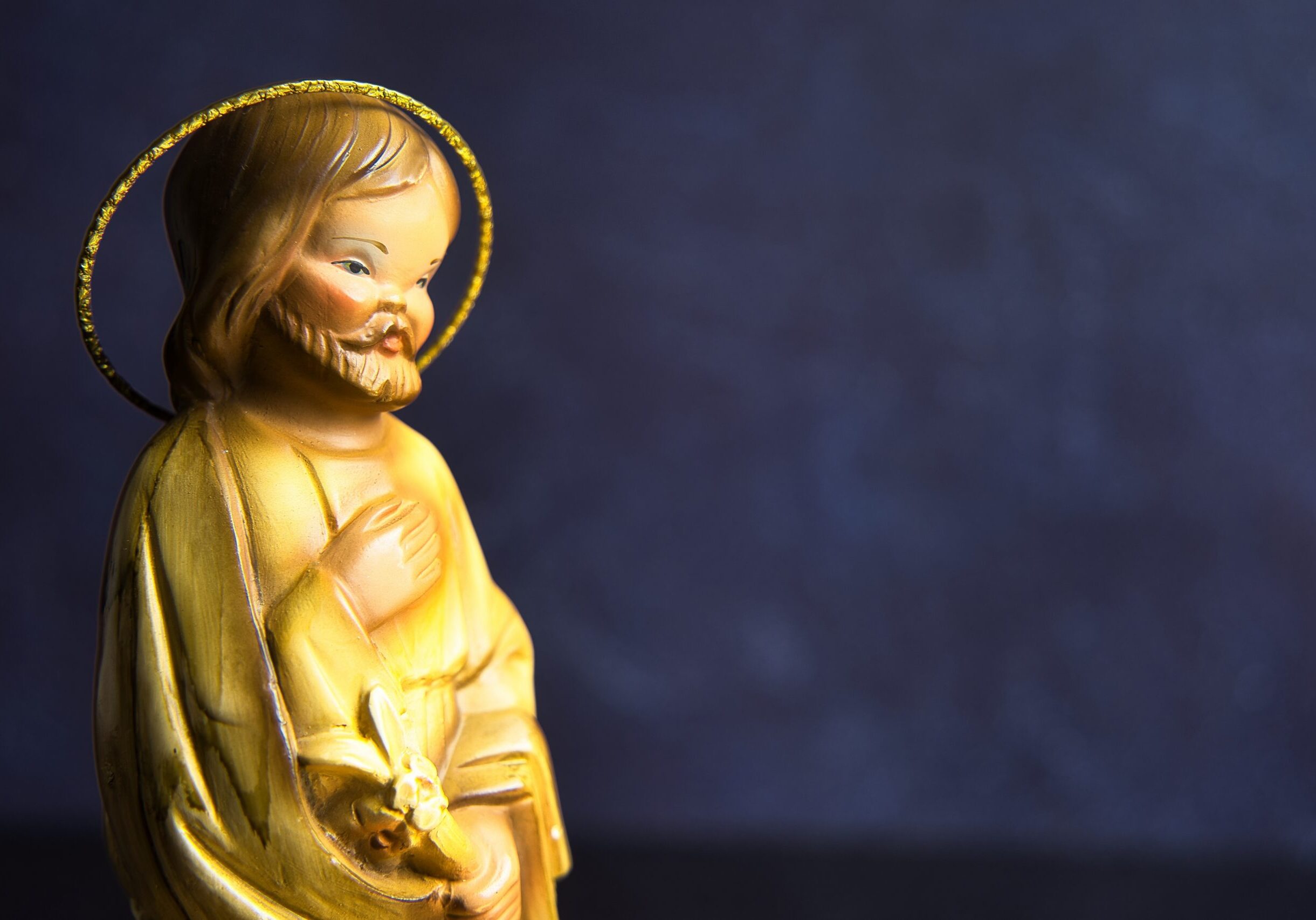 A small golden statue of Saint Joseph with a halo, shown in profile against a dark blue background, holding a lily and looking downward with a gentle expression.