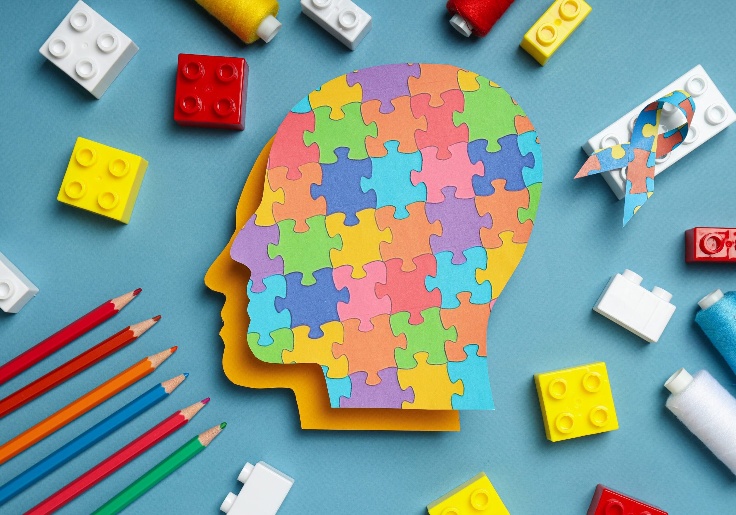 A colorful profile of a human head made of puzzle pieces sits on a blue background, surrounded by crayons, building blocks, and thread, symbolizing autism, creativity, and different ways of thinking.