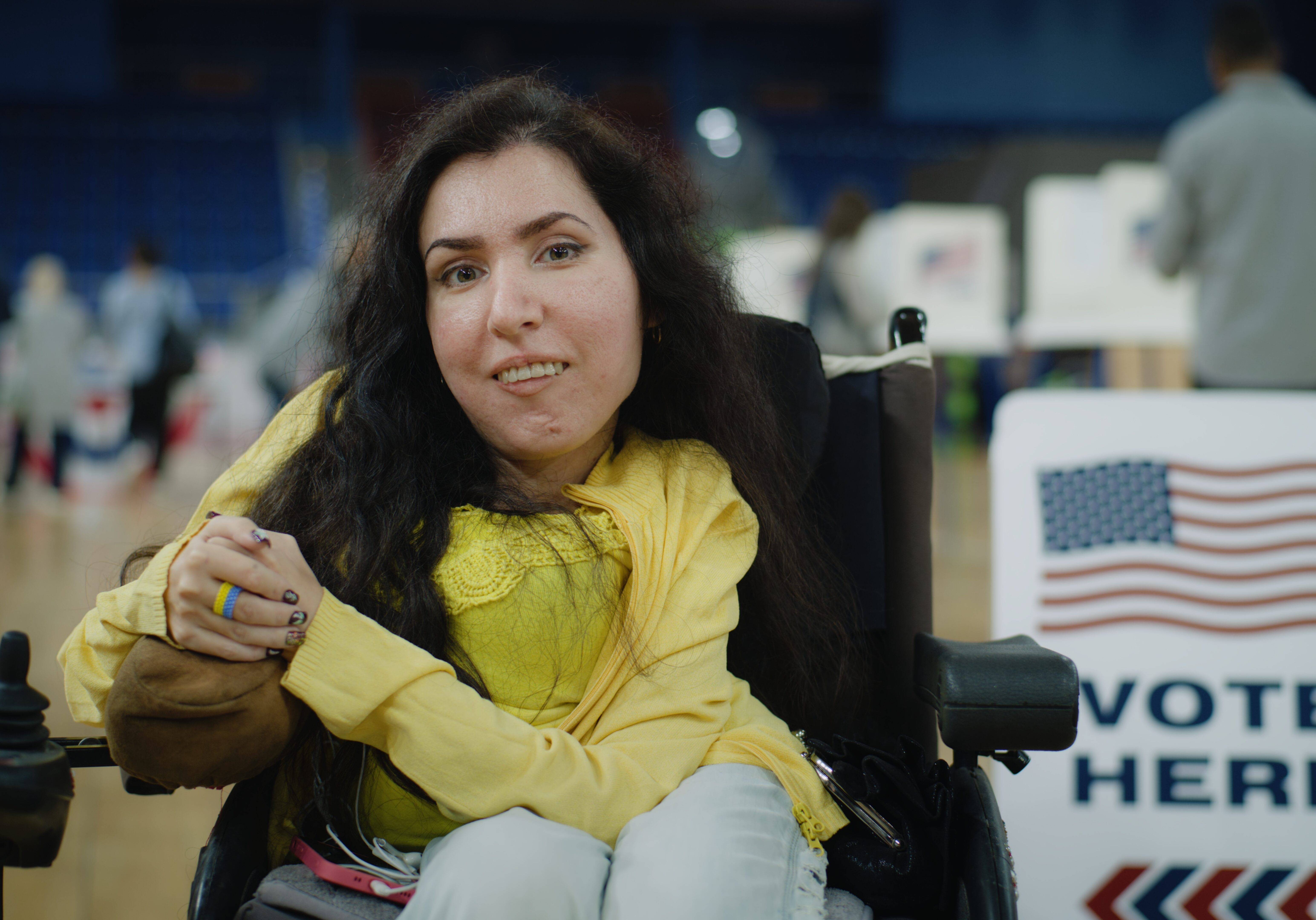 A woman in a wheelchair smiles at the camera inside a polling place. She is wearing a yellow top and sitting near a “Vote Here” sign decorated with an American flag. Other voters are blurred in the background as they cast their ballots.