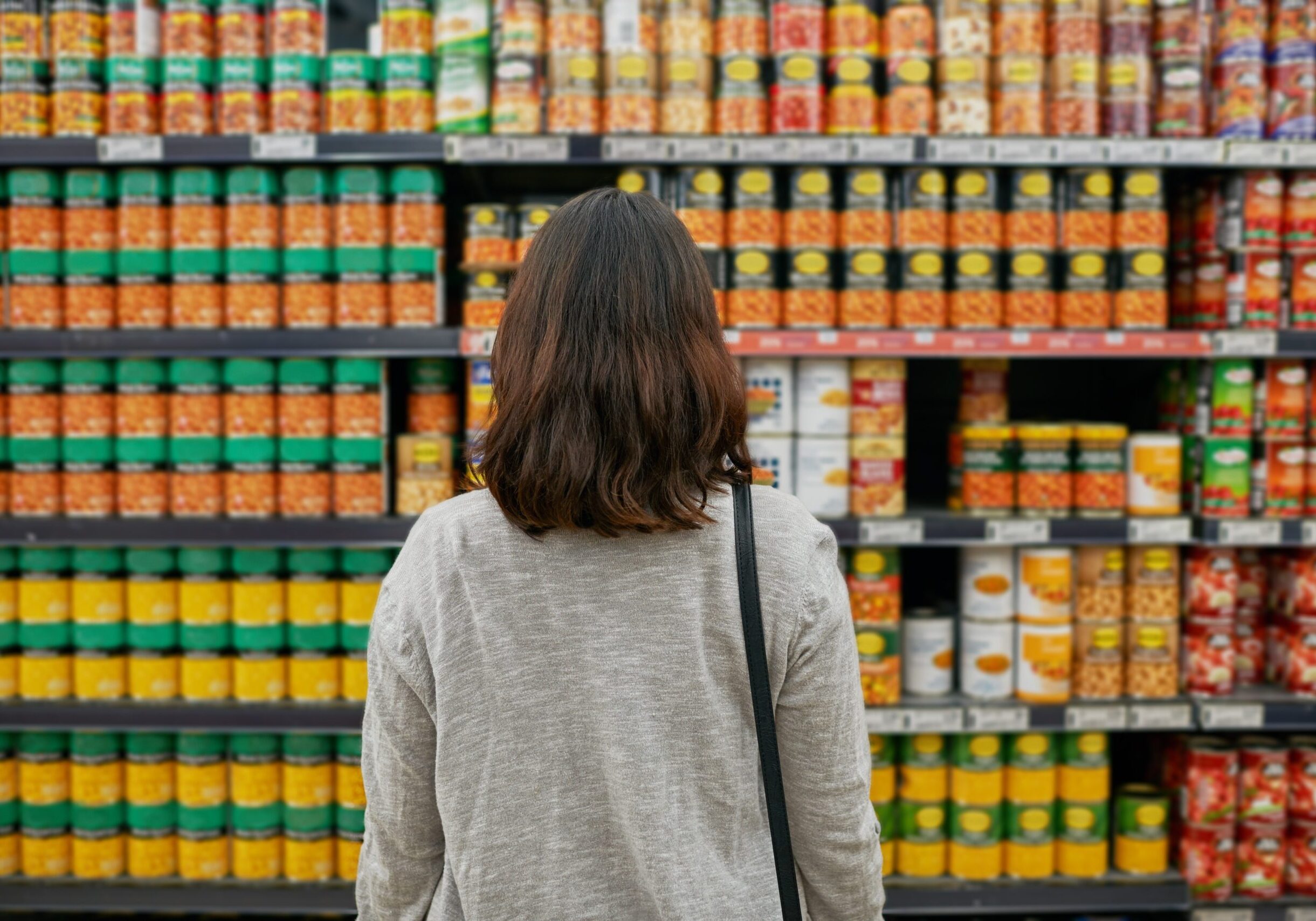 A woman stands in front of shelves of canned food, reflecting on the growing cost of groceries and the reality of hunger after SNAP benefits were cut.