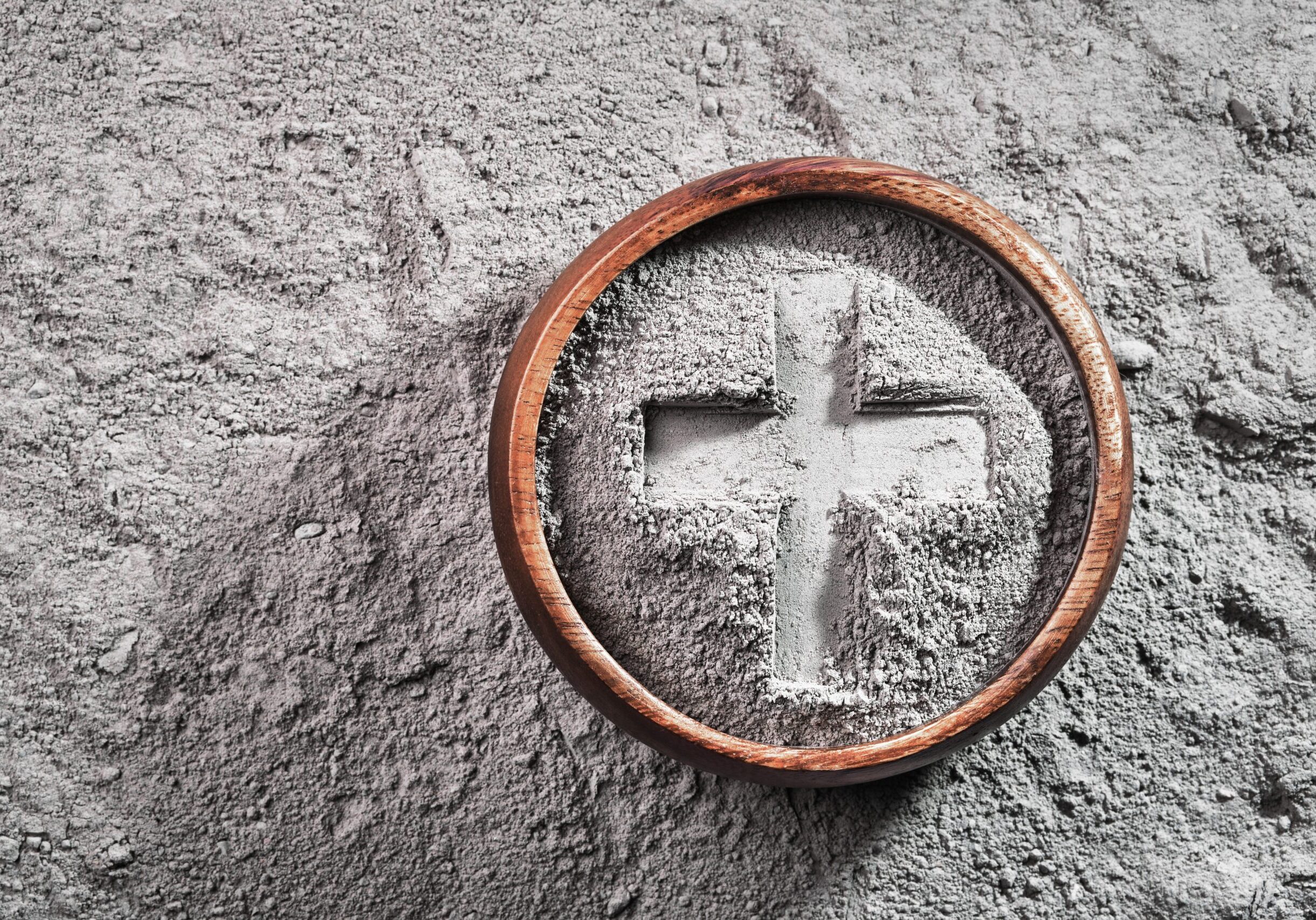 Close-up of gray ash arranged in the shape of a cross inside a round wooden bowl, resting on a textured gray surface.