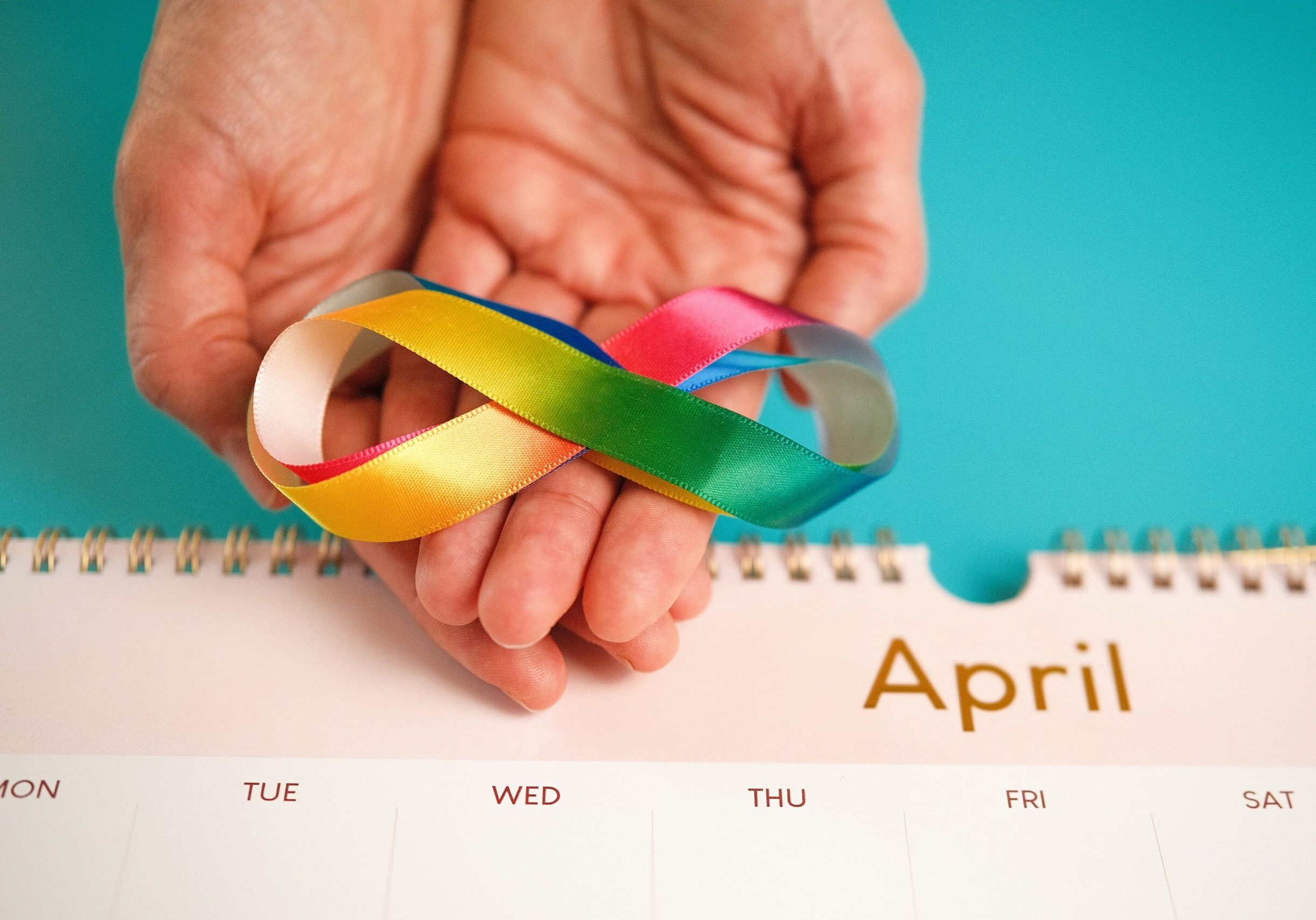 A hand holds a rainbow infinity ribbon above a calendar page labeled April, representing Autism Awareness Month and the beauty of neurodiversity.