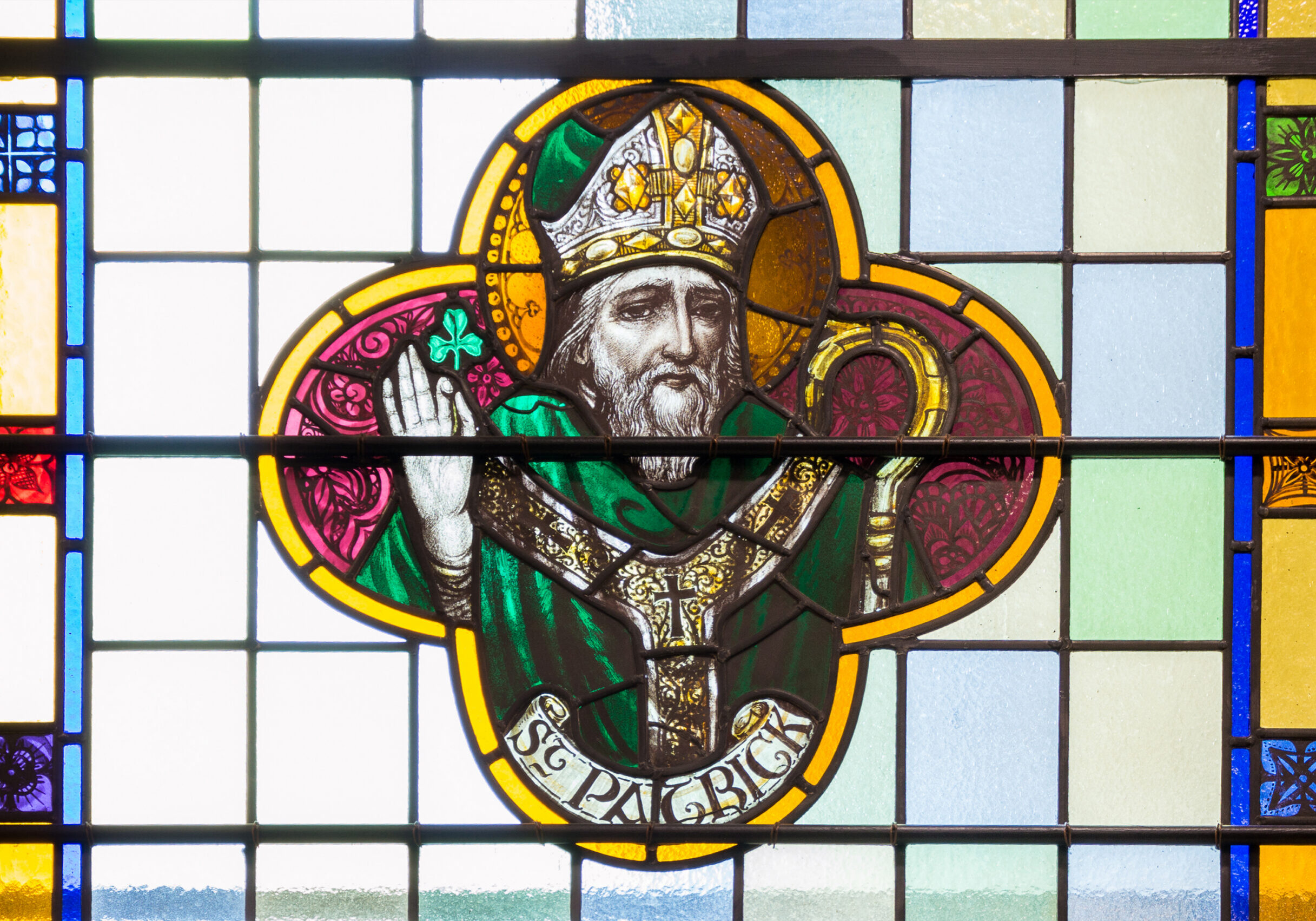Stained glass window depicting Saint Patrick, shown wearing a bishop’s mitre and green robes, raising one hand in blessing while holding a crozier, framed by a clover-shaped design and colorful geometric panes in the background.