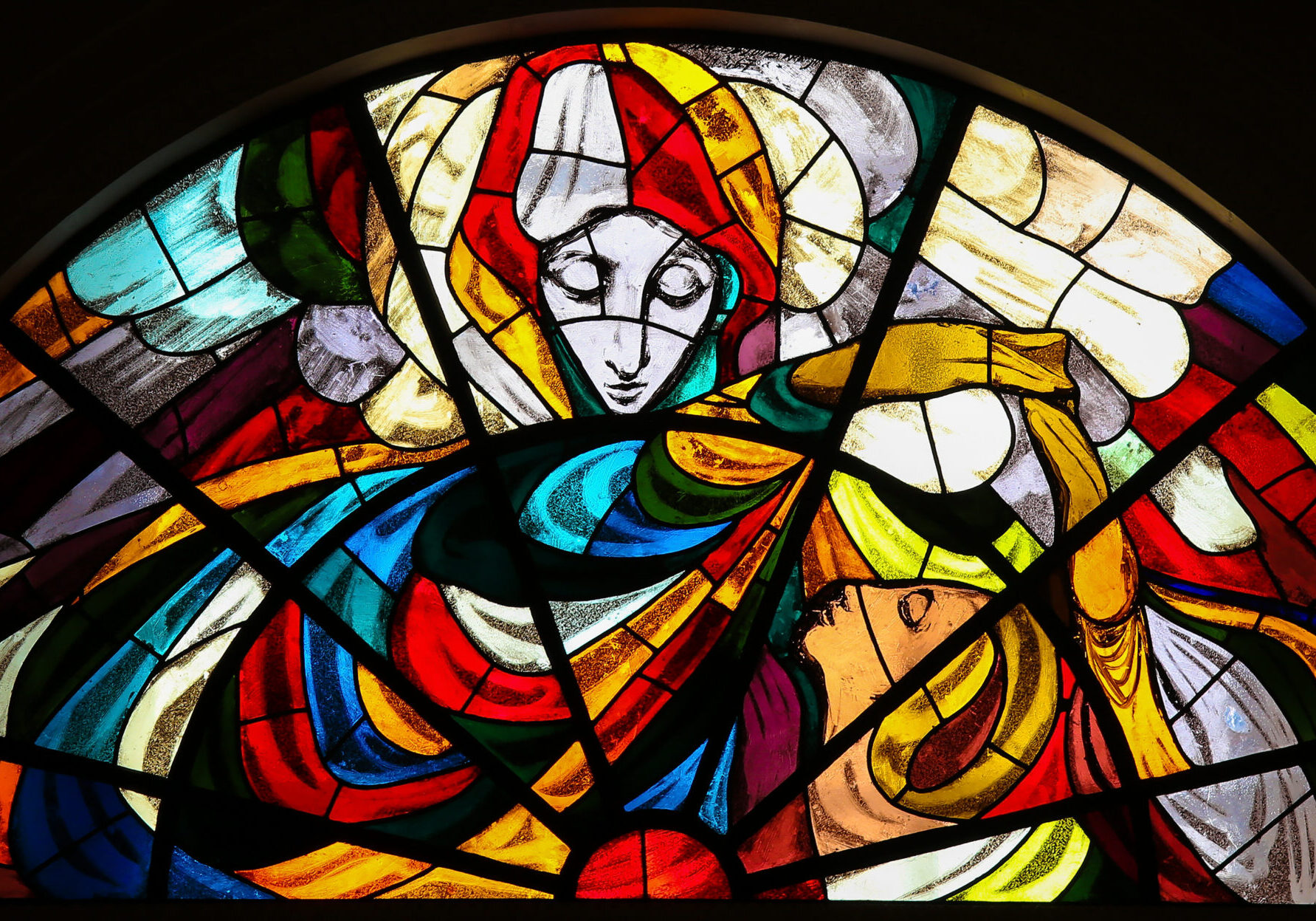 A colorful stained-glass window depicting a serene, Blessed Mother in flowing robes leaning toward another person who gazes upward, bathed in radiant light, with vivid reds, blues, yellows, and greens forming an abstract, sacred scene suggestive of a biblical encounter.