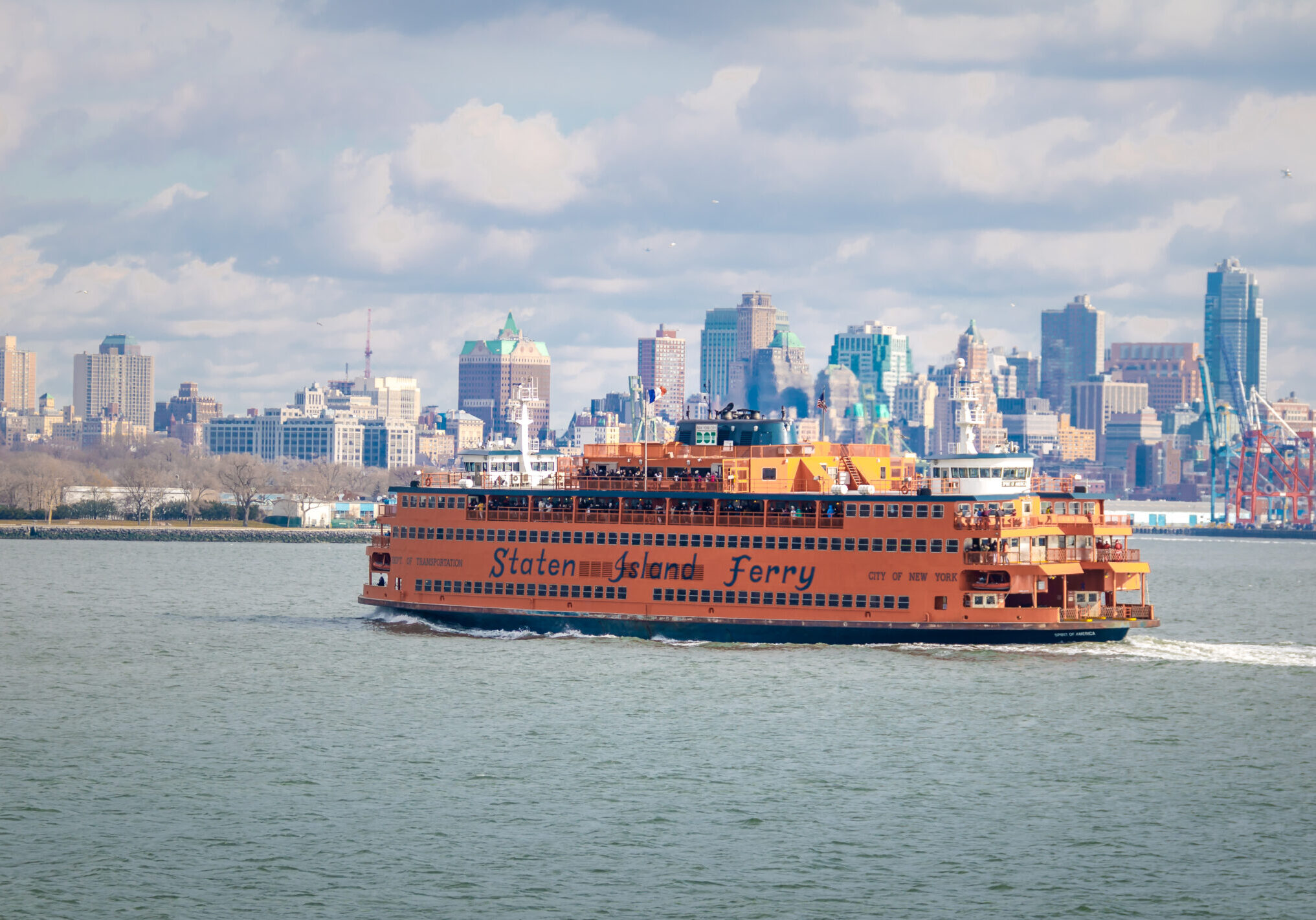 A bright orange Staten Island Ferry sails across New York Harbor with the Manhattan skyline in the background under a partly cloudy sky.